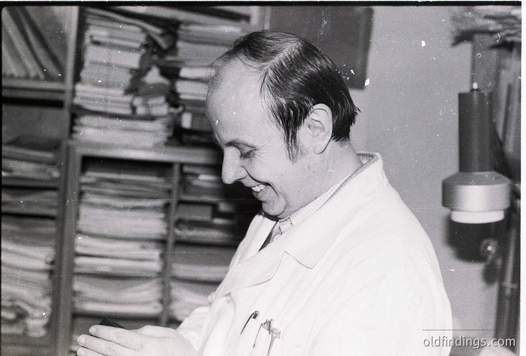 Mid-20th century black-and-white photo of a man in a lab coat examining rolled microfilm or film reels in a storage cabinet. Industrial archive setting, likely 1950s–1970s.