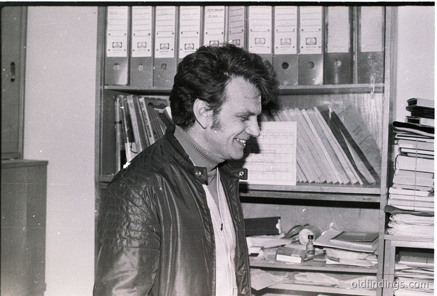 Mid-century office setting featuring a man in a leather jacket and collared shirt, smiling at the camera. Surrounding him are metal shelving units filled with binders, files, and stacked documents. The scene suggests a 1960s–1970s administrative or academic environment.