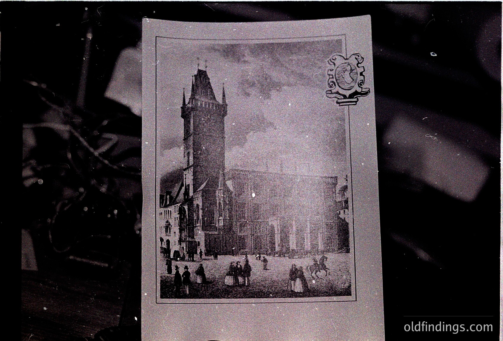 Vintage postcard featuring a Gothic-style tower with crenellated parapets and a clock, likely part of a European cathedral or town hall. Crowds of 19th-century attire gather in a courtyard below. Art Nouveau-style embossed seal in top-right corner.