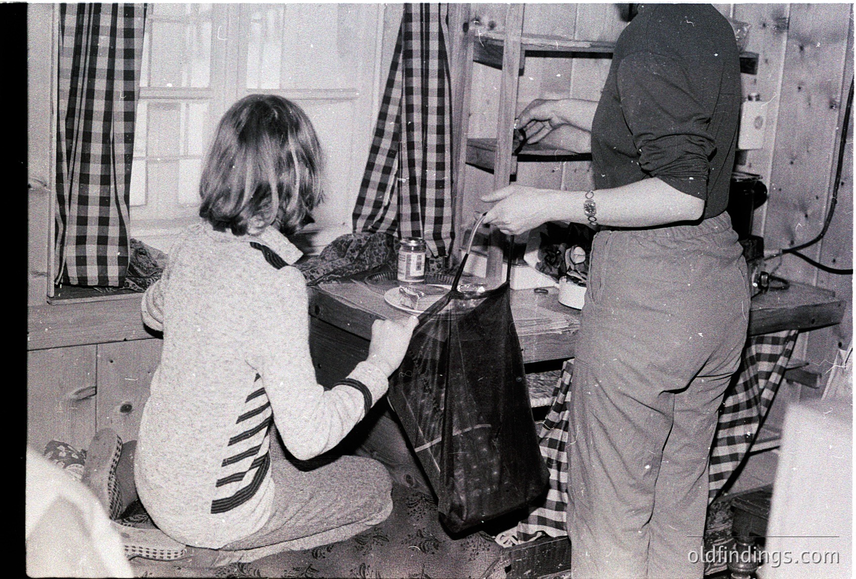 Two women in a mid-century kitchen, likely 1960s–1970s, preparing food. One kneels, holding a jar, while the other stands, pouring from a can into a pot on a wooden table. Checkered curtains and a black stove with a kettle suggest domestic life. Rustic, functional decor hints at post-war European home life.