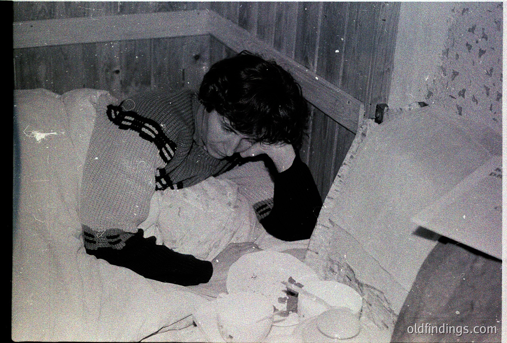 Black-and-white photo of a person sleeping on a mattress in a damaged, partially collapsed room. Exposed bricks and debris suggest structural damage, likely from conflict or natural disaster. Clothing appears worn, indicating possible wartime or post-war conditions. Mid-20th century setting, possibly Eastern Europe.