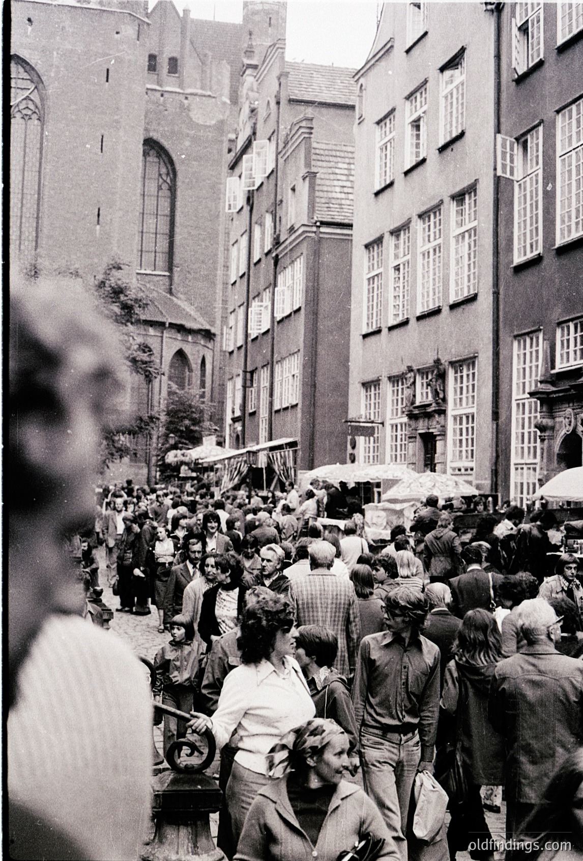 Crowded European street market in 1960s-70s, flanked by brick buildings with arched windows. Stalls line the narrow alley, selling goods under makeshift canopies. Mid-century fashion and utilitarian architecture dominate.