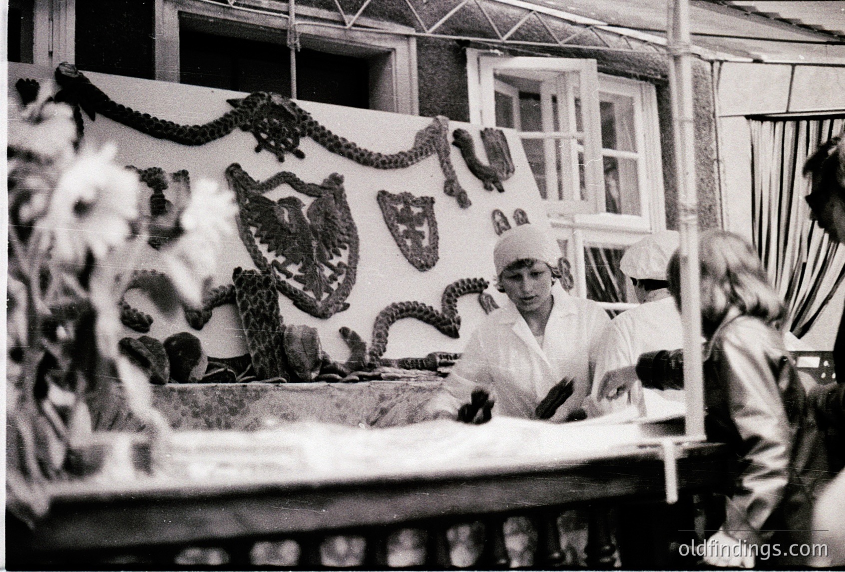 Industrial-era food processing facility featuring workers in white uniforms and hairnets. Central figure handles large, coiled seafood (likely squid) on a conveyor belt. Decorative wall plaques with maritime motifs (anchor, fish) suggest a coastal or fishing-themed workspace. Mid-20th century (1950s–1970s) industrial design with bright overhead lighting.