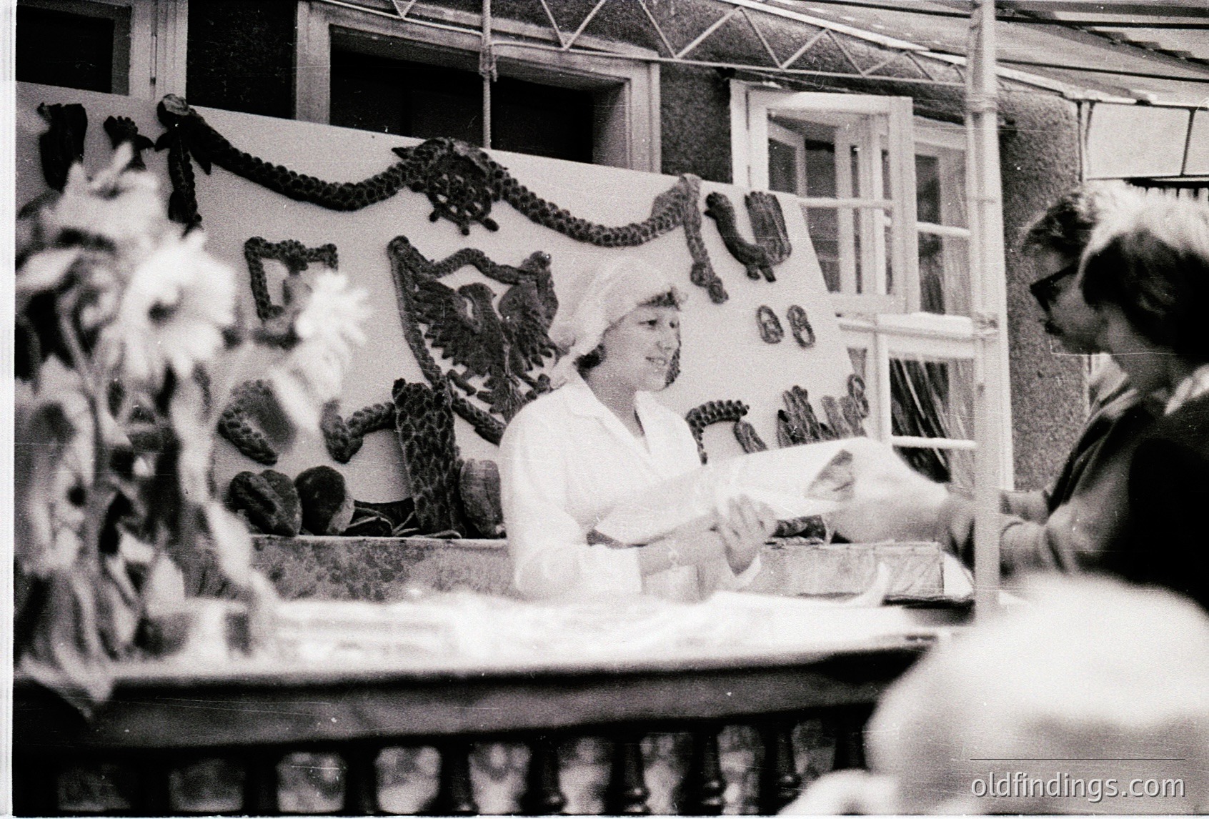 Vintage black-and-white photo of a woman in a white chef’s uniform and hat preparing food at a public market stall, likely 1960s–1970s. Decorative metal eagle sculpture and hanging meat cuts in background. Indoor market setting with wooden counter and shelving.