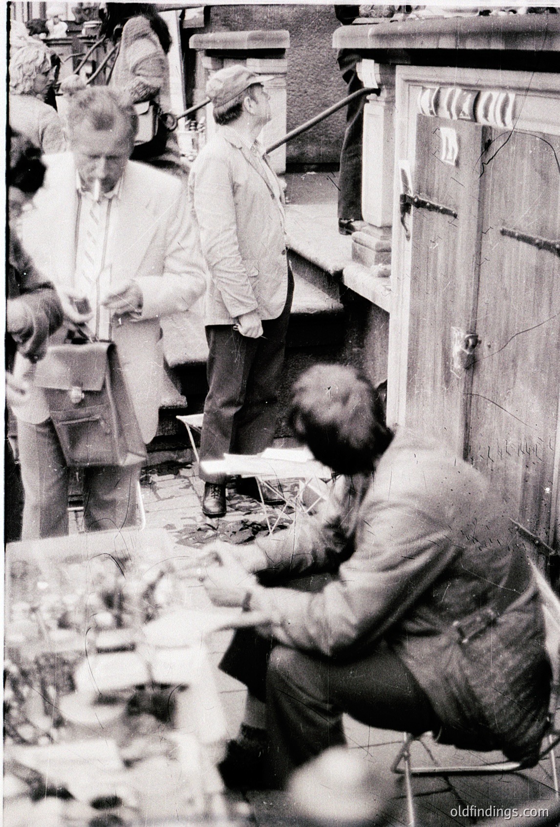 Vintage black-and-white photo of three men engaged in detailed craftwork in a dimly lit workshop. One man sits at a table with tools and materials, while others stand or crouch, focusing on intricate tasks. Industrial-era setting with visible machinery and wooden workbenches. Likely late 19th to early 20th century, possibly a watchmaker’s or clockmaker’s workshop.