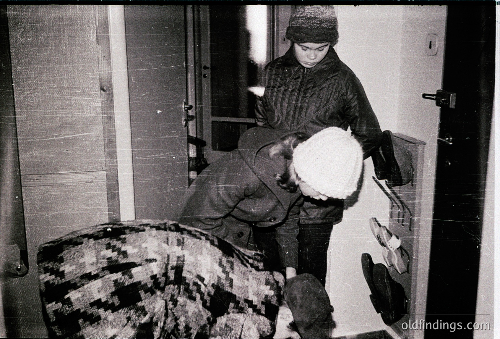 Mid-20th century indoor scene: A child in winter attire (hat, coat, gloves) carries a large white fur coat over their shoulder while stepping through a doorway. A patterned rug lies on the floor, and a pair of boots rests nearby. Simple interior with wooden door and wall paneling.