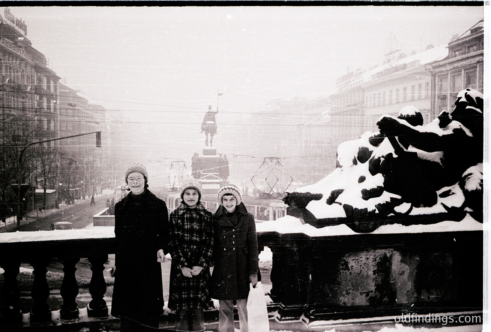 Where is this: three children pose on a snow-covered railing in a winter cityscape, likely mid-20th century