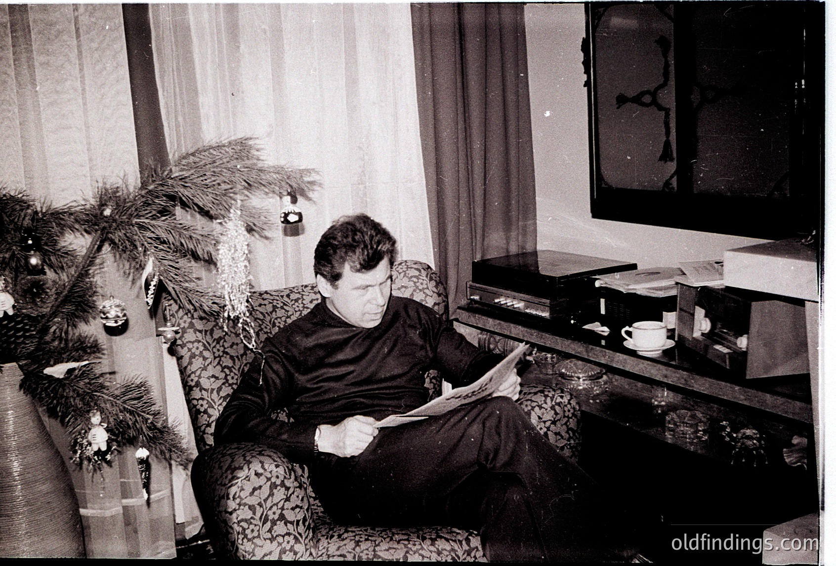 Mid-century living room scene: man in formalwear reads newspaper beside a decorated Christmas tree. Vintage TV console holds a teacup, ashtray, and records. Floral-patterned armchair and curtains suggest 1960s–1970s domestic interior. écor