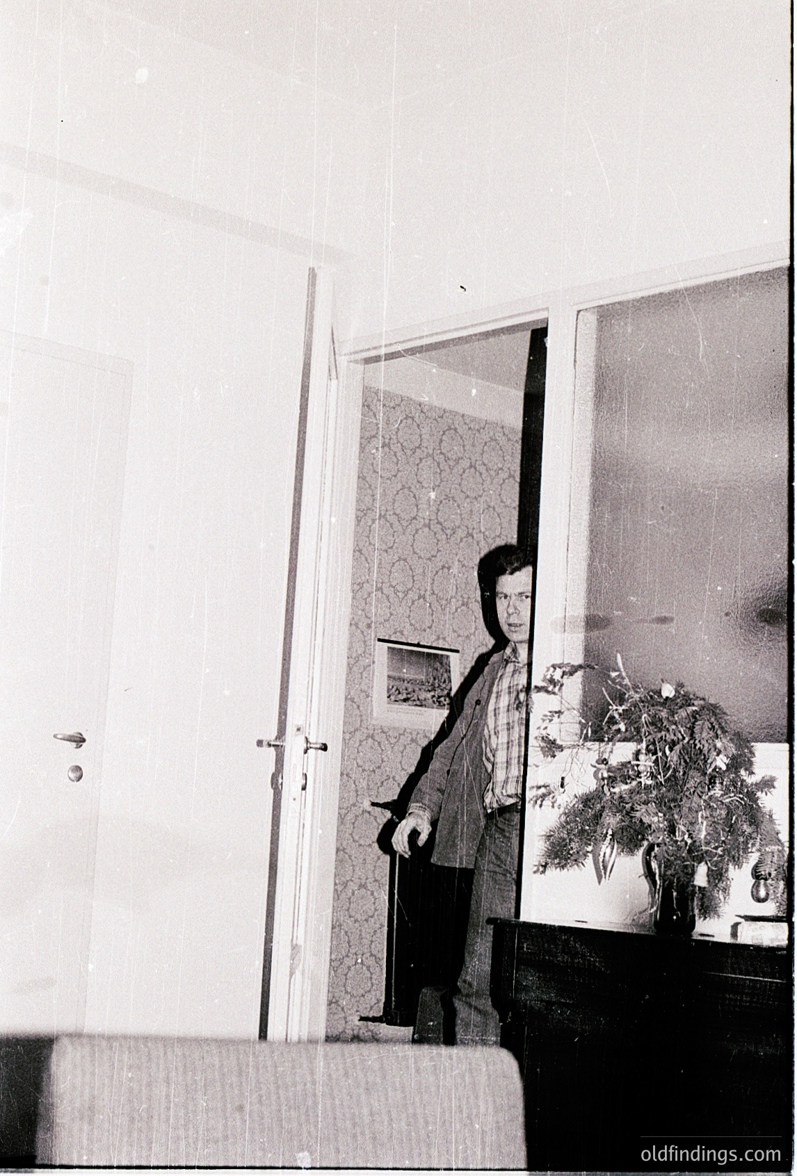 Mid-century indoor portrait: person in doorway holding a framed photo, framed mirror above, floral-patterned wallpaper. Likely 1960s–1970s domestic interior.