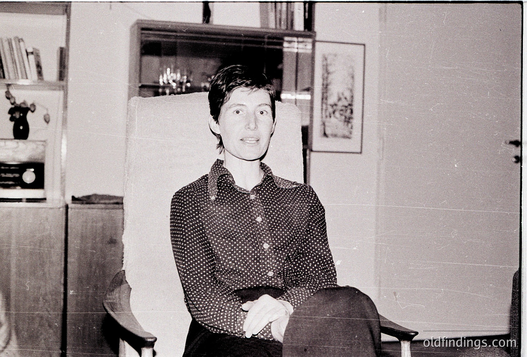 Black-and-white portrait of a person in a patterned blouse seated indoors, likely mid-20th century. Bookshelves and framed artwork in a minimalist office or study setting.