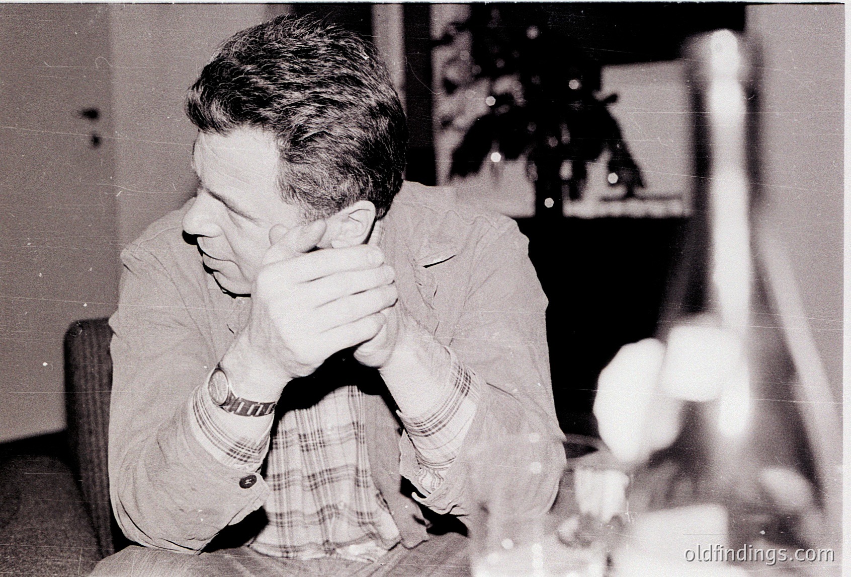 Man in mid-20th-century indoor setting, covering face with hands. Wearing checkered shirt, wristwatch, and seated near a table with glassware. Indoor lighting suggests evening gathering. Style hints at or Western Europe/US.