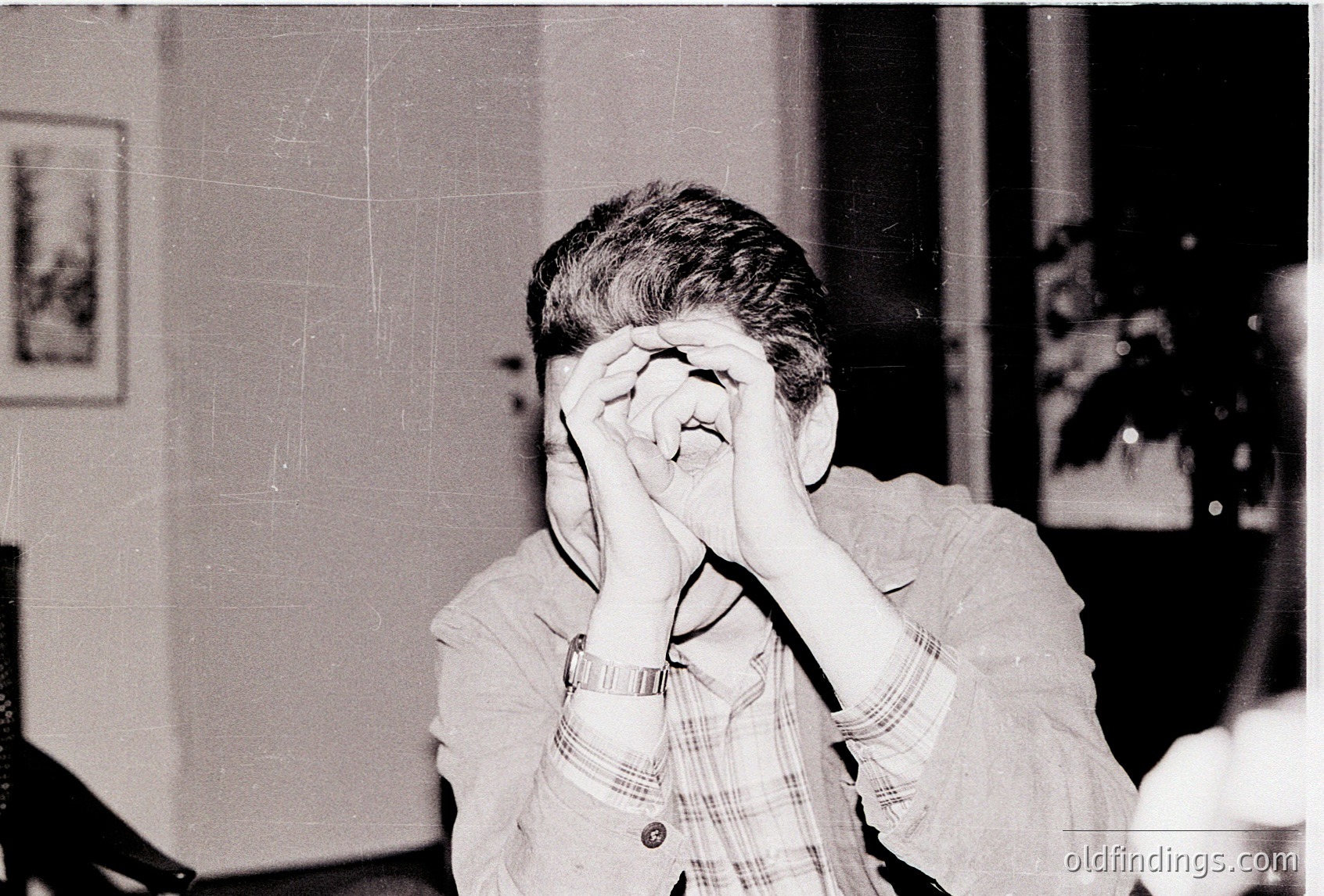 Black-and-white candid of an individual covering their face with hands, seated indoors. Wearing a checkered shirt with a wristwatch, likely mid-20th century. Background shows framed art, a potted plant, and a blurred interior setting.
