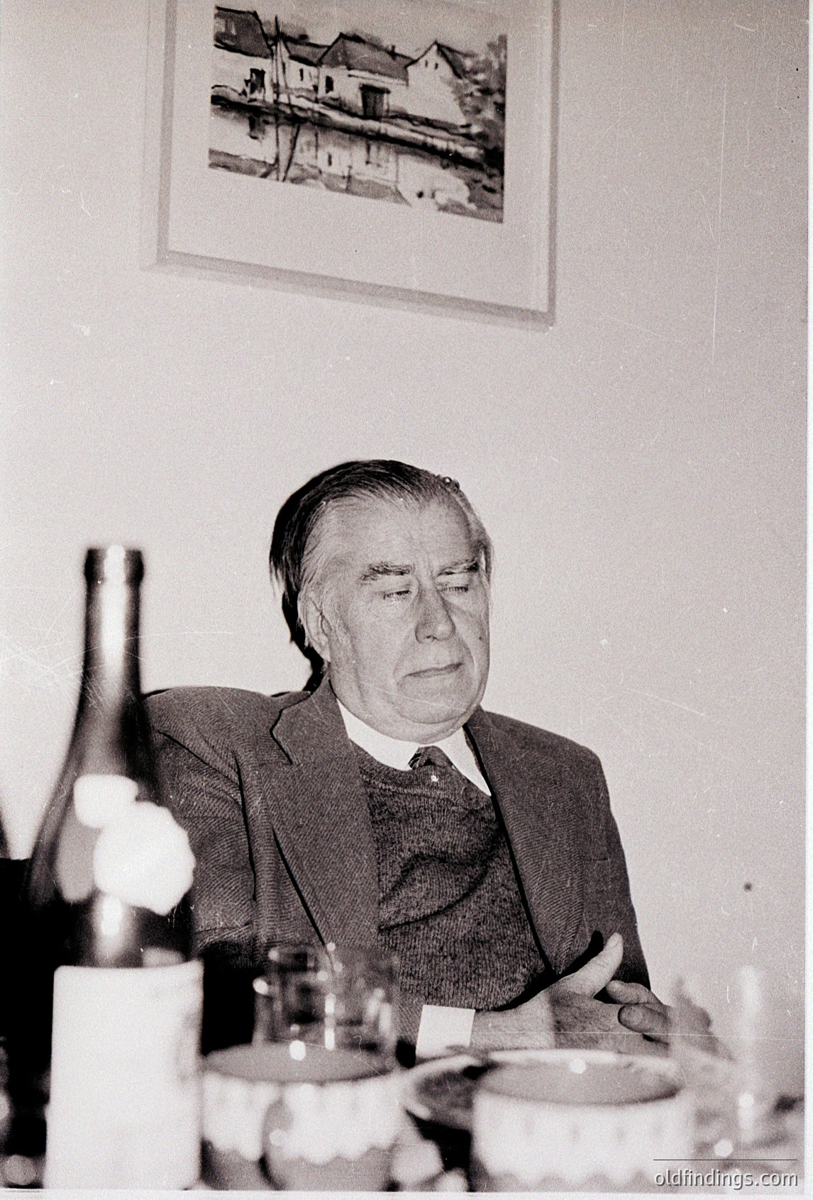 Mid-20th century portrait of a man in formal attire, seated at a table with wine bottles and glasses. Above him, a framed landscape print depicts a rural scene with a river and houses. Style suggests 1950s–1960s Western European or North American setting.
