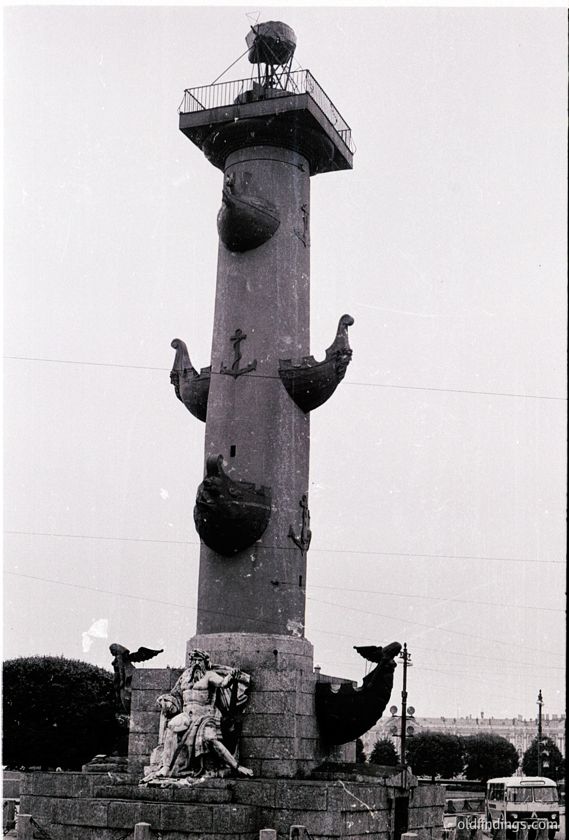 Tall stone column with maritime sculptures: a central figure at base holding a shield, flanked by winged sea creatures and anchors. Top features a platform with a spherical object. Likely a 19th-century maritime monument.