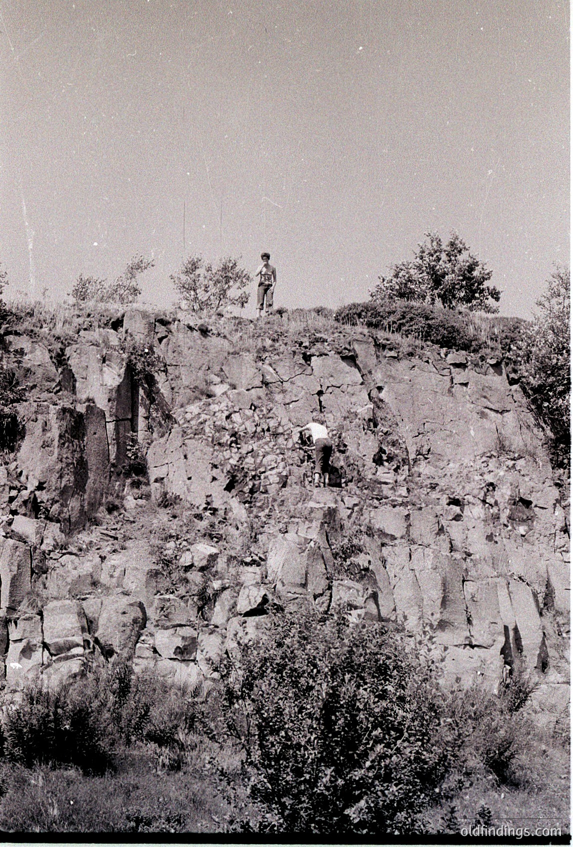 Vintage black-and-white photo of rugged rocky terrain with two figures—one standing atop a cliff, one descending via a narrow path. Dense shrubbery and exposed rock layers dominate the scene. Likely mid-20th century, possibly for hiking or geological study.