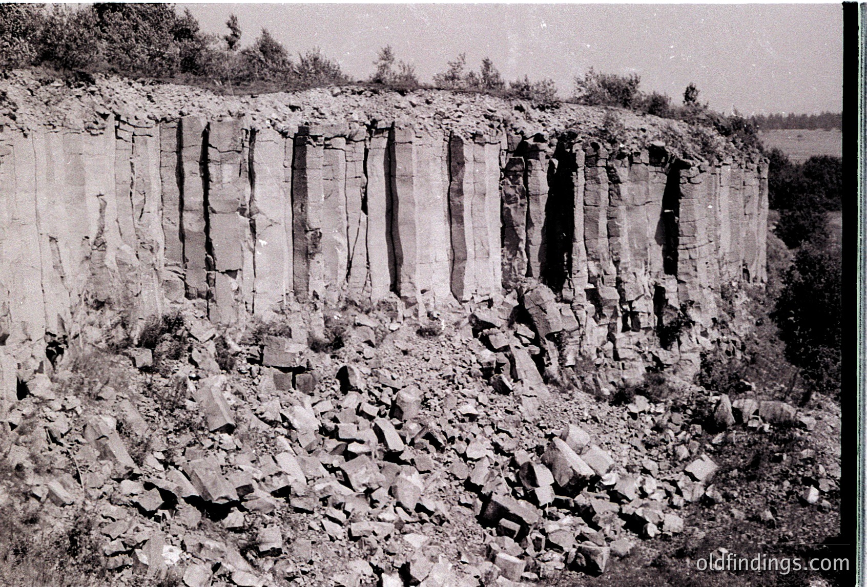 Vertical limestone quarry face with stacked, rectangular blocks and debris at base, mid-20th century industrial site. Distinctive stratified rock layers visible in cross-section.