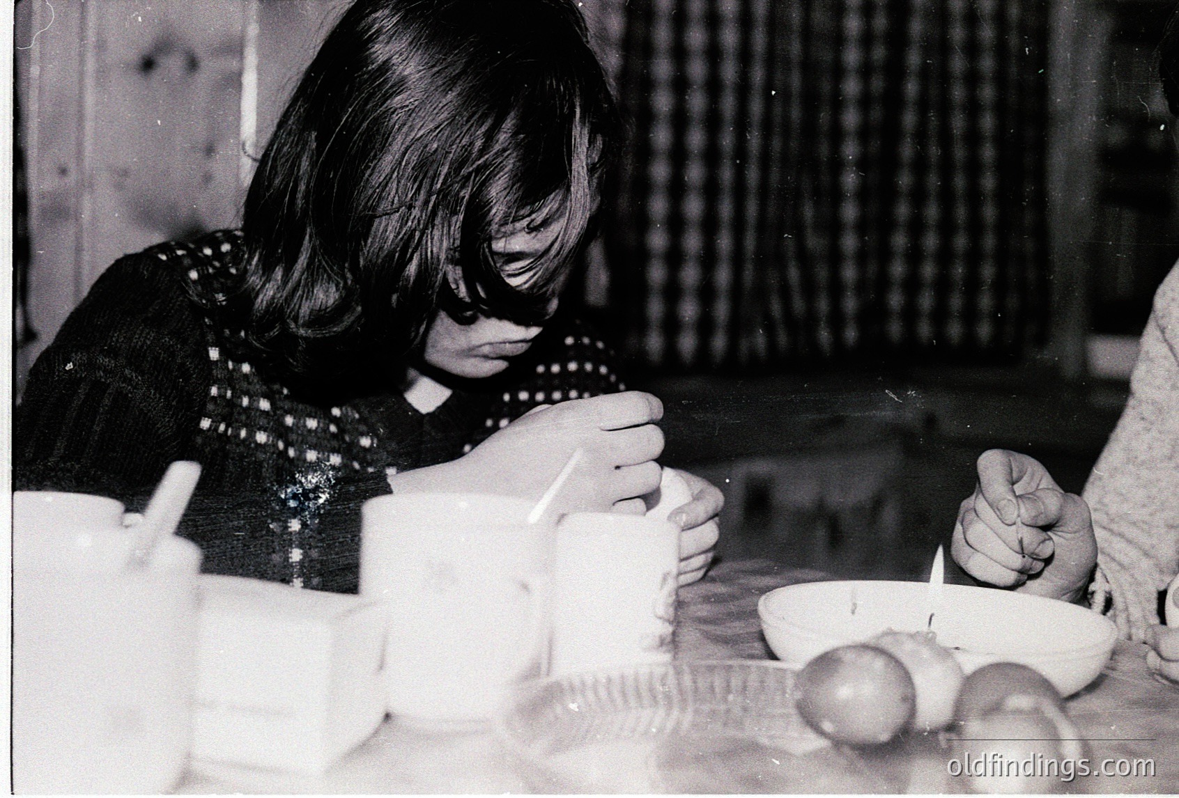 Vintage black-and-white photo of two individuals seated at a rustic wooden table, engaged in a meal. The person on the left wears a patterned sweater and holds a spoon, while a bowl of food and a glass of drink are visible. The setting suggests a cozy, domestic atmosphere, likely mid-20th century.