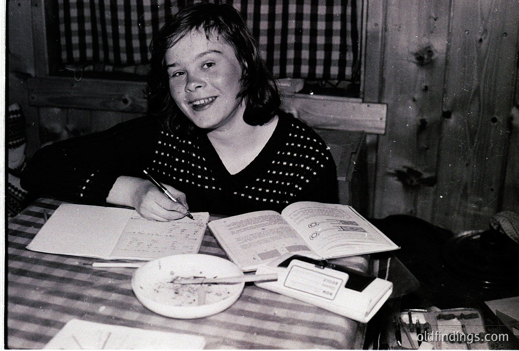 Young girl writing at a wooden table in a rustic indoor setting, likely mid-20th century. She wears a polka-dot sweater and holds a pen over an open booklet with stamps. A plate of cookies and a small notebook sit nearby.