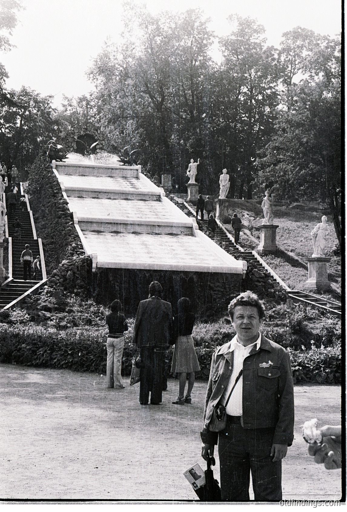 Mid-20th century black-and-white photo of a man in a military-style jacket posing in a grand park setting. Behind him, a symmetrical staircase flanked by statues leads to a tiered, terraced structure. Lush greenery and mature trees frame the scene. Visible 1960s-70s fashion and architecture suggest a European urban park.