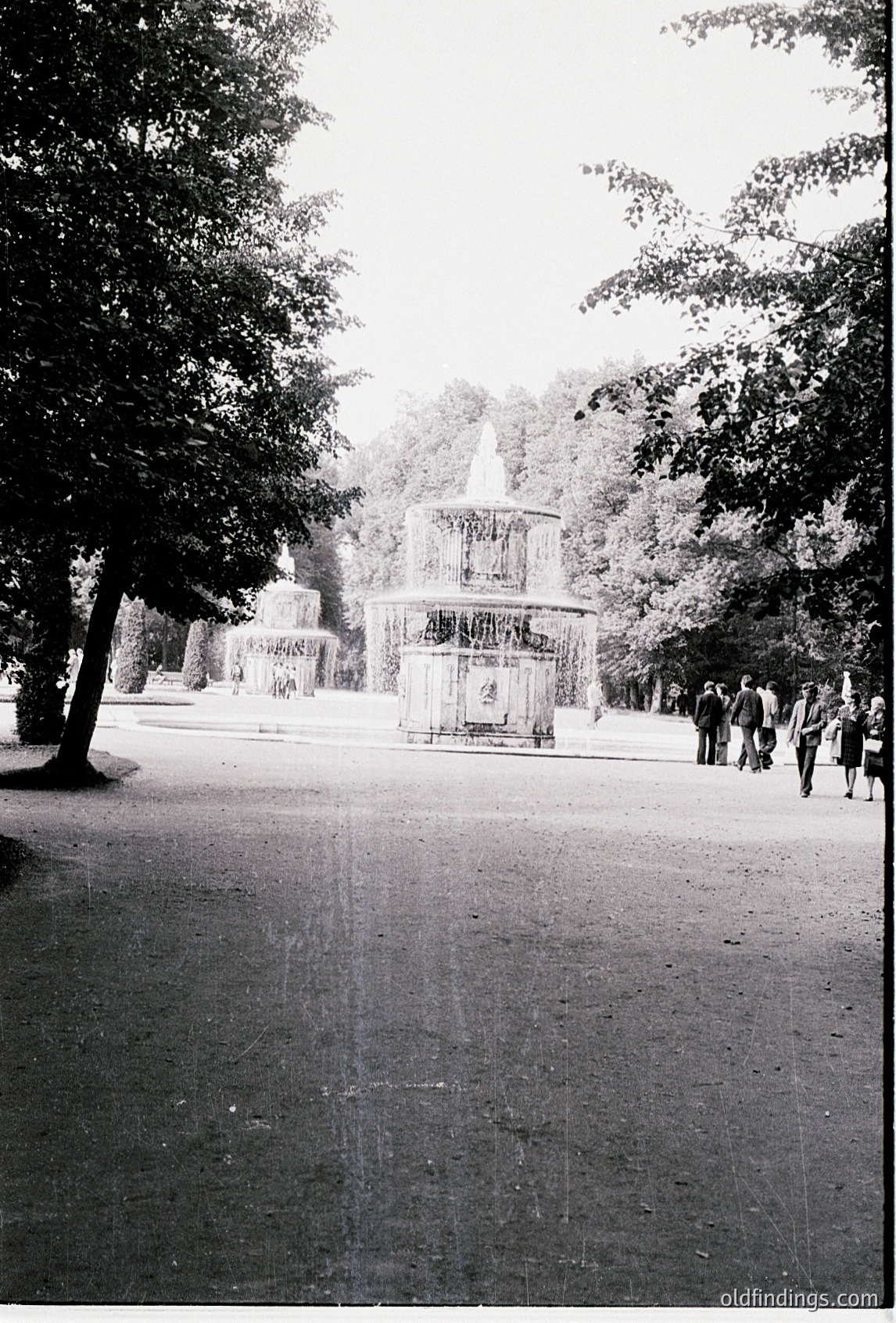 Neoclassical fountain with ornate ironwork and central water spout in a tree-lined park pathway, mid-20th century. Group of people in formal attire (suits, dresses) walking toward the structure, suggesting a public or ceremonial event.