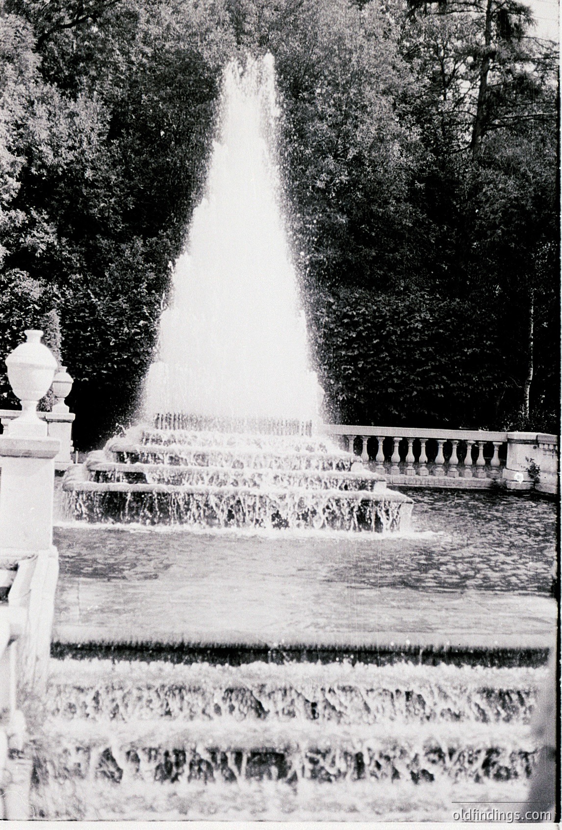 Grand Baroque-style fountain with cascading tiers, framed by dense greenery. Symmetrical design features ornate balustrades and Latin inscriptions. Likely European palace or estate garden, 17th–18th century.