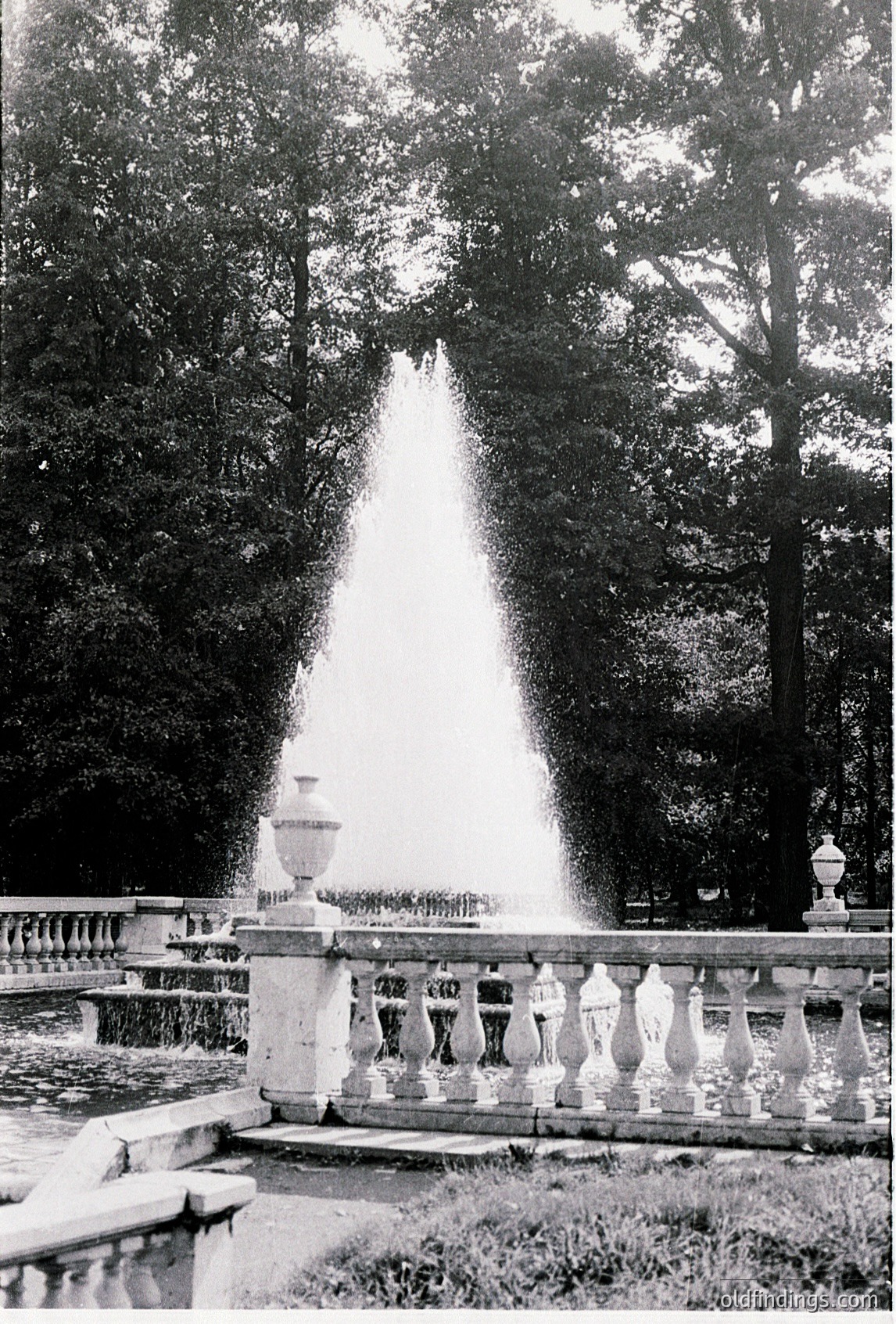 Classic 19th-century fountain with symmetrical stone balustrade and tall, conical water jet framed by mature trees. Likely European park or estate design, showcasing neoclassical landscaping.