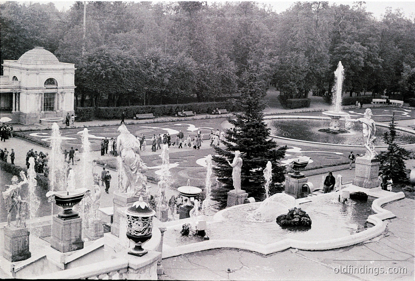 Grand classical fountain complex with ornate sculptures and cascading water in a public park. Neoclassical pavilion in background. Mid-20th century European urban design.