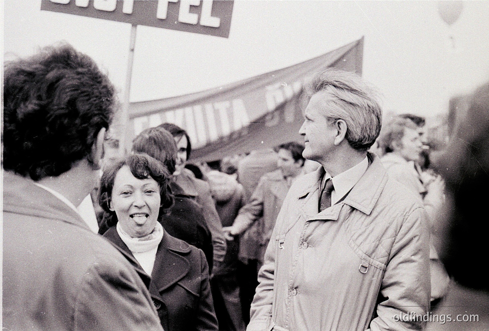 Black-and-white protest scene featuring two central figures in mid-conversation, surrounded by a crowd. The man wears a light jacket with a pin, the woman a collared blouse. A visible sign reads "SIGTETEL" (likely a reference to Sighet, Romania). Crowd density and 1970s-era clothing suggest Eastern Bloc activism. Crowd dynamics and signage hint at political demonstration.