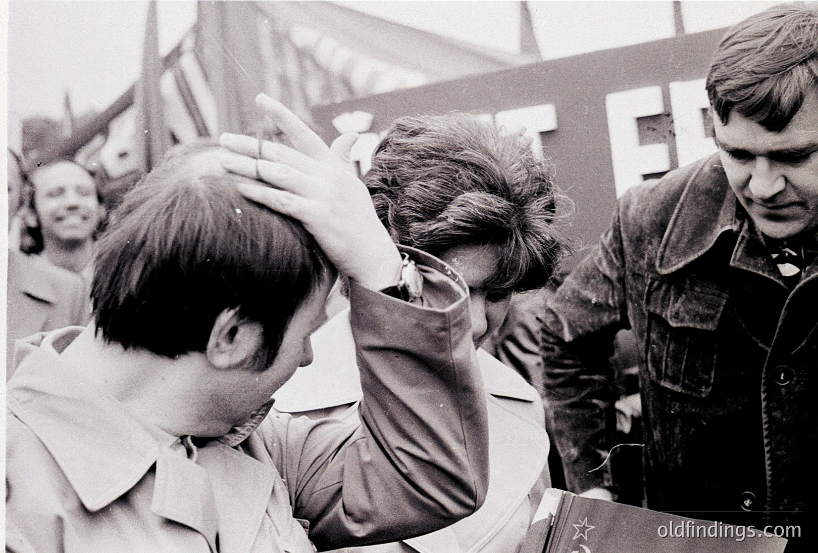 Intimate black-and-white moment of three men in close emotional exchange, likely 1960s–1970s Eastern Bloc. Foreground figure raises hand to another’s face, while a third observes with arms crossed. Military-style jackets and badges suggest political or labor context. Urban setting with indistinct buildings and blurred crowd in background.