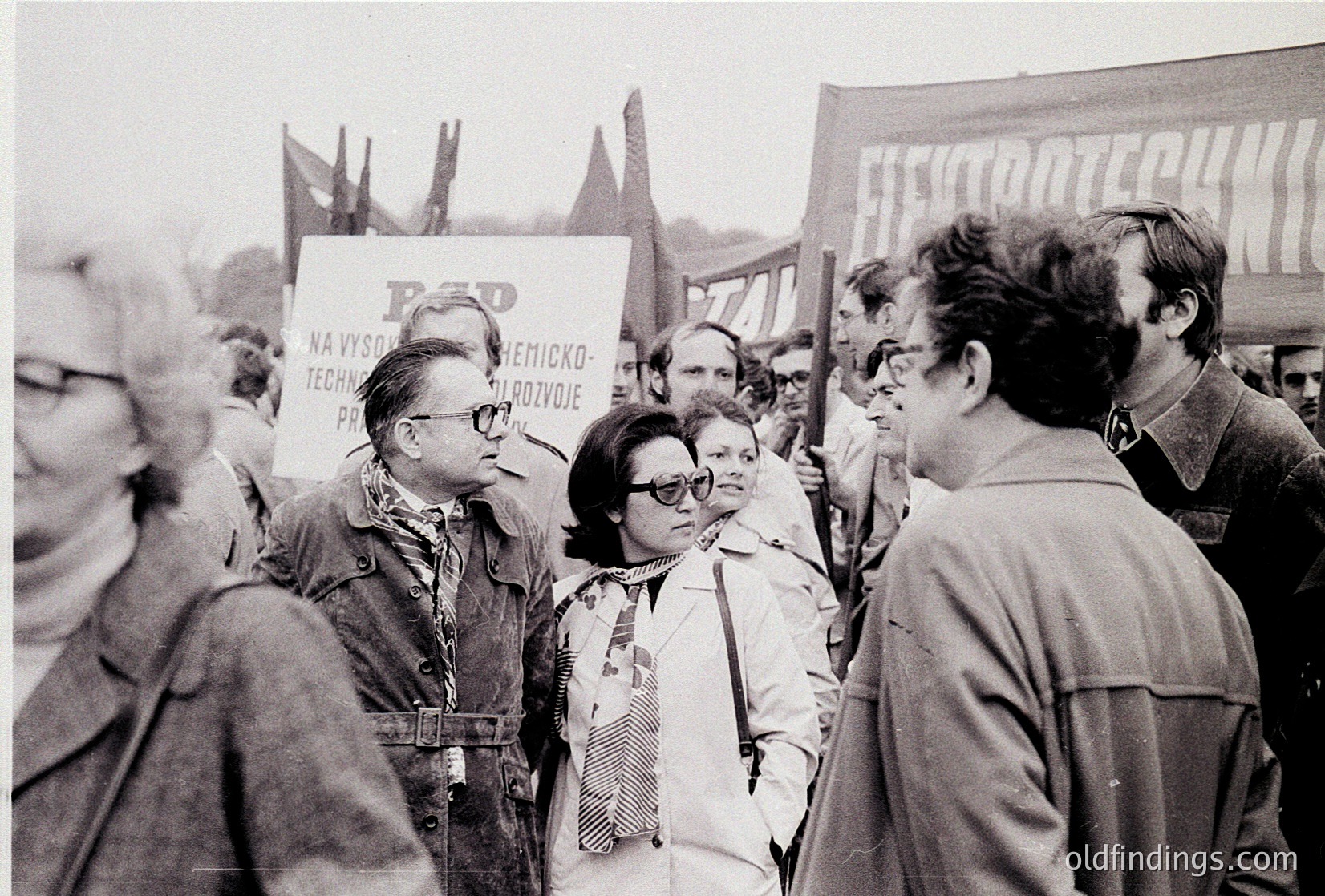 Black-and-white protest scene featuring a crowd holding signs with Cyrillic text, likely Eastern Bloc era. Central figures wear 1970s-80s fashion—men in jackets, women in patterned blouses. Prominent sign reads "BĚP" with chemical/industrial text, suggesting a labor or environmental rally. Urban setting with indistinct industrial backdrop.