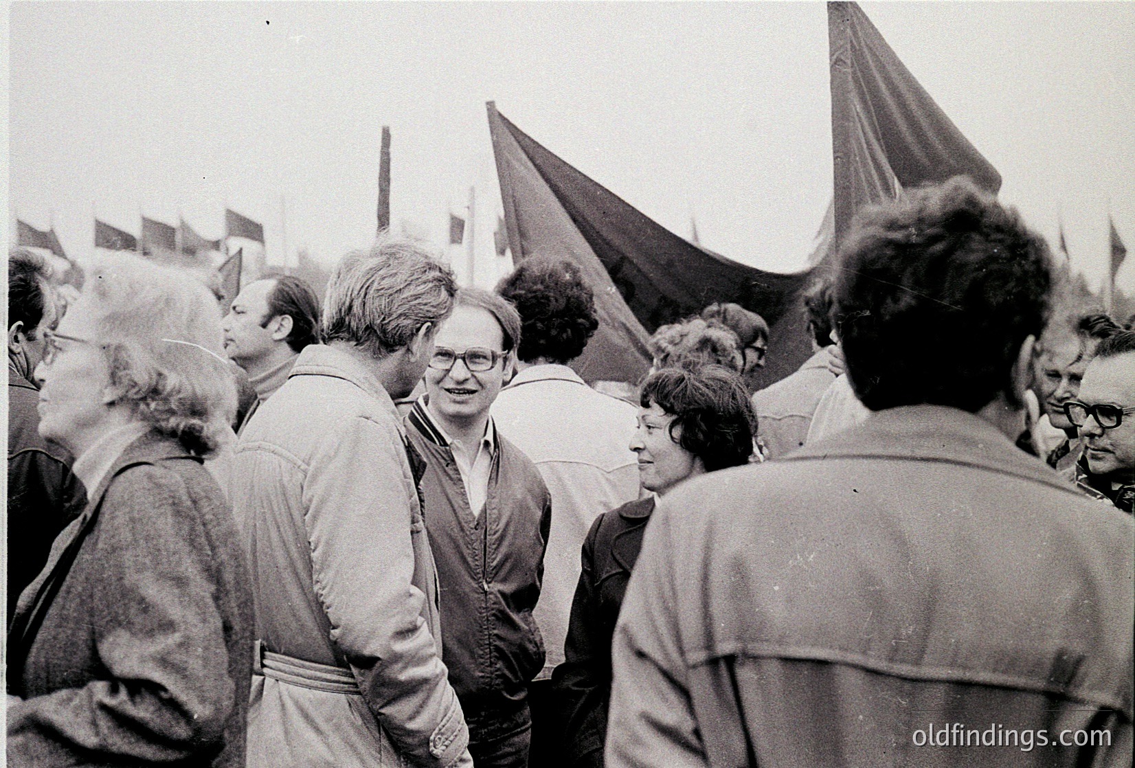 Black-and-white photo of a mid-20th-century public gathering, likely 1960s–1970s. Crowd of adults in casual attire (jackets, sweaters) with some wearing glasses, engaged in conversation. Prominent black flags with white symbols in background suggest a political or labor demonstration. Urban setting with indistinct buildings.