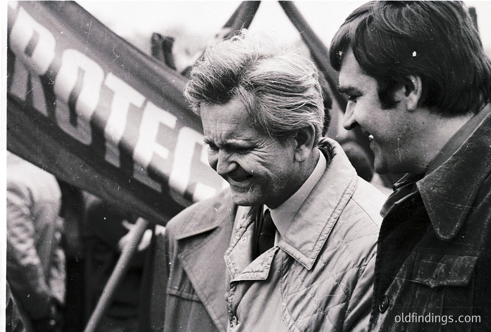 Black-and-white photo of two men at a public event, likely 1960s–1970s. The man in focus wears a light jacket and appears engaged in conversation, with a banner reading "ROTER" (German for "red") in the background, suggesting a political rally or demonstration. Crowd and microphones indicate a speech or gathering.