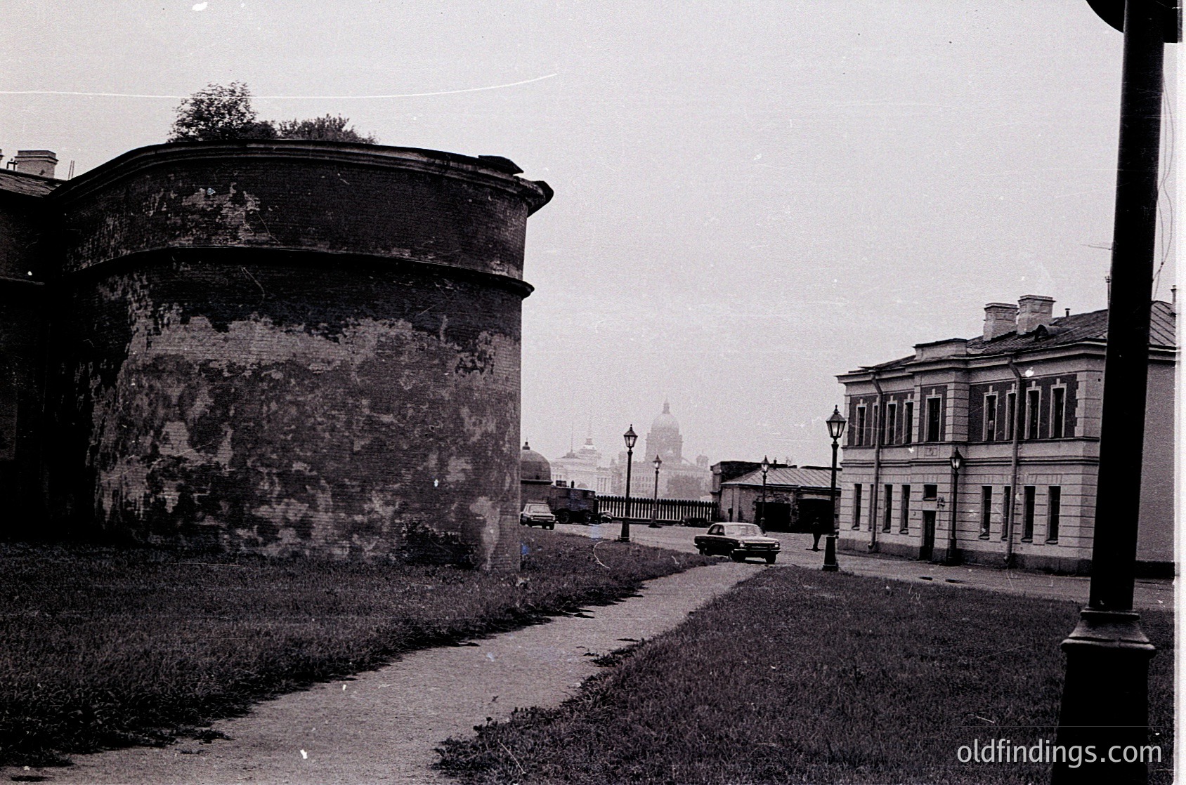 Vintage black-and-white street scene featuring Soviet-era architecture. Prominent round fortress wall with weathered plaster, flanked by a neoclassical building with decorative columns. A single vintage car drives along a paved path toward a distant bridge and domed church spire. Overcast sky enhances the historic atmosphere.