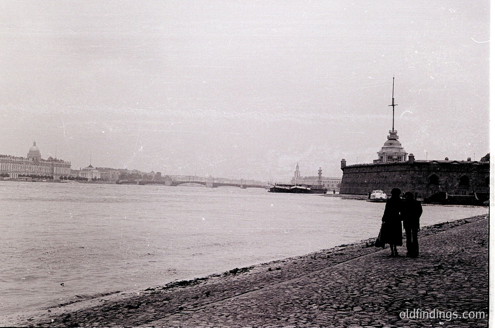 Black-and-white view of St. Petersburg’s Neva River promenade, featuring the Peter and Paul Fortress and its spire in the foreground. Two figures walk along the cobblestone riverside, with the Kryukov Bridge and Winter Palace complex visible in the background. Early 20th-century urban landscape with maritime architecture.