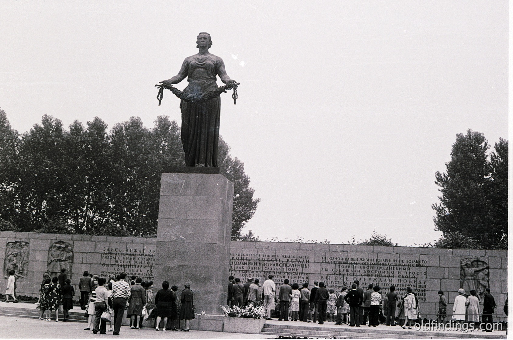 Monumental statue of a robed figure holding a scroll, standing atop a stone pedestal with inscribed plaques. Crowd gathers in solemn procession, some placing floral tributes. Soviet-era memorial design, likely –1970s. --- *Note: The inscribed text and architectural style suggest a Soviet-influenced war memorial, potentially from Bulgaria’s communist period.*