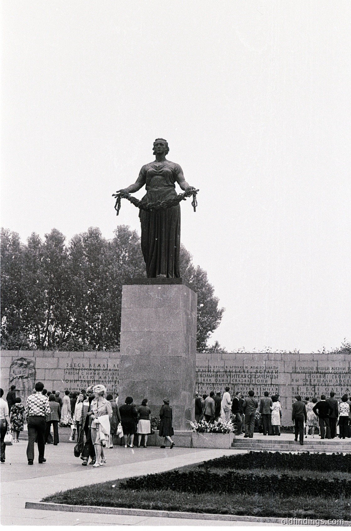 Monumental statue of a robed woman holding a wreath atop a stone pedestal, surrounded by engraved plaques. Crowd of people in 1960s-era clothing gathers in a public square, likely a memorial site.