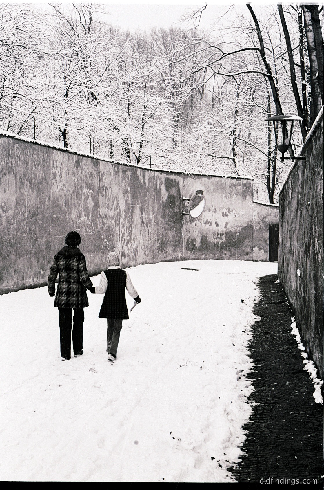 Two figures walk hand-in-hand along a snow-covered path beside a high concrete wall, framed by bare winter trees. The woman wears a patterned coat; the child, a light jacket. Graffiti of a bird appears on the wall. Likely urban winter scene, mid-20th century.