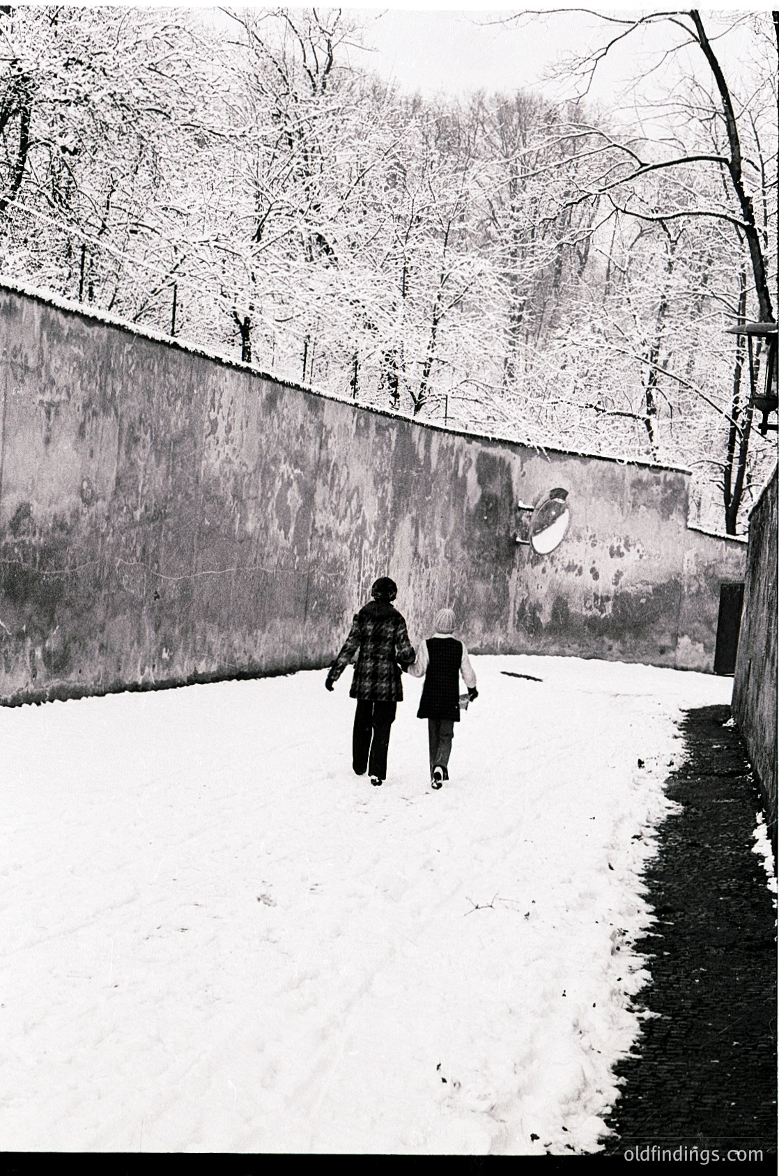 Two children walk hand-in-hand on a snow-covered path beside a concrete wall, framed by bare winter trees. Mid-20th century urban setting, likely Eastern Europe.
