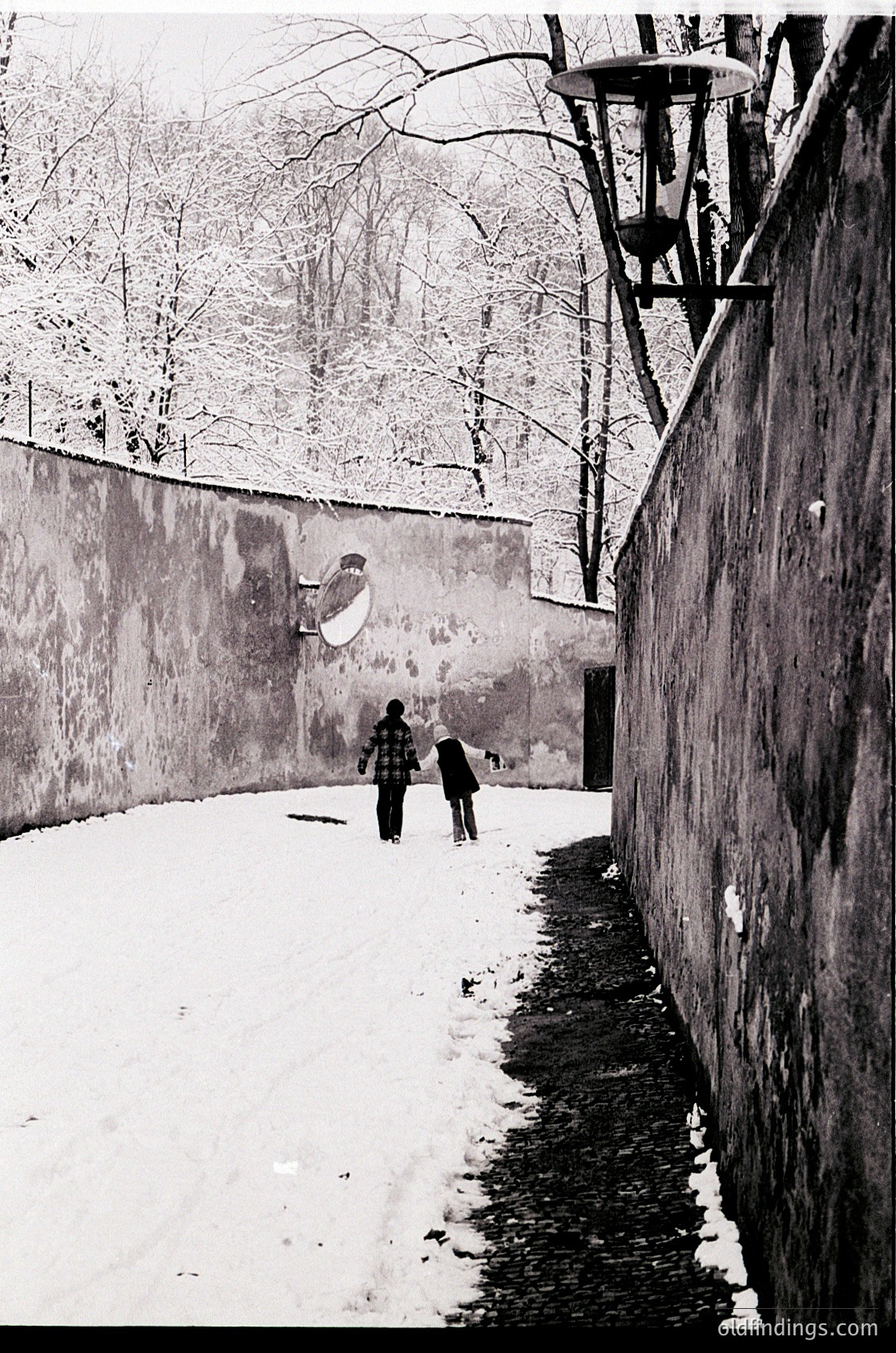 Black-and-white street scene featuring two figures walking along a snow-covered alley flanked by high walls. Snow accumulates on bare branches and rooftops, suggesting winter. A vintage streetlamp and graffiti-like mural on the wall add urban texture. Likely Eastern European urban setting, mid-20th century.