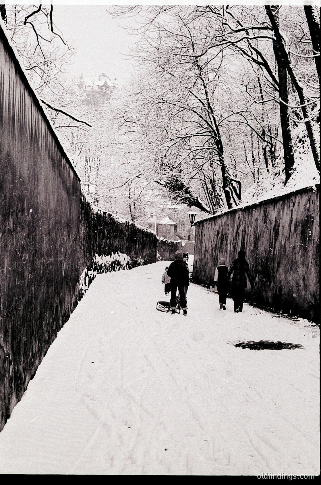Black-and-white street scene featuring three children sledding down a snow-covered path flanked by high concrete walls. Bare winter trees and distant buildings frame the urban winter landscape.