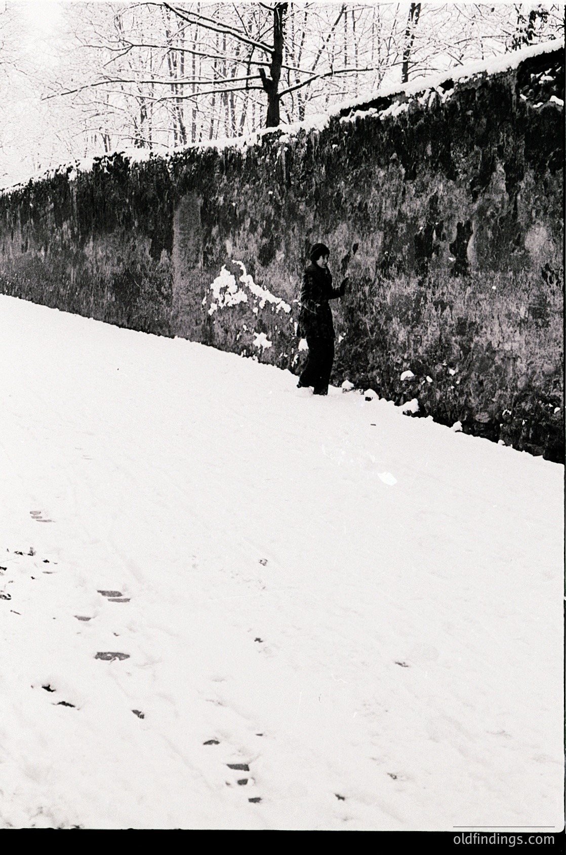 Black-and-white street scene featuring a lone figure in winter attire—long coat, hat, and gloves—standing beside a stone wall. Snow blankets the ground and rooftops, with bare trees in the background. Likely urban setting, early-to-mid 20th century.