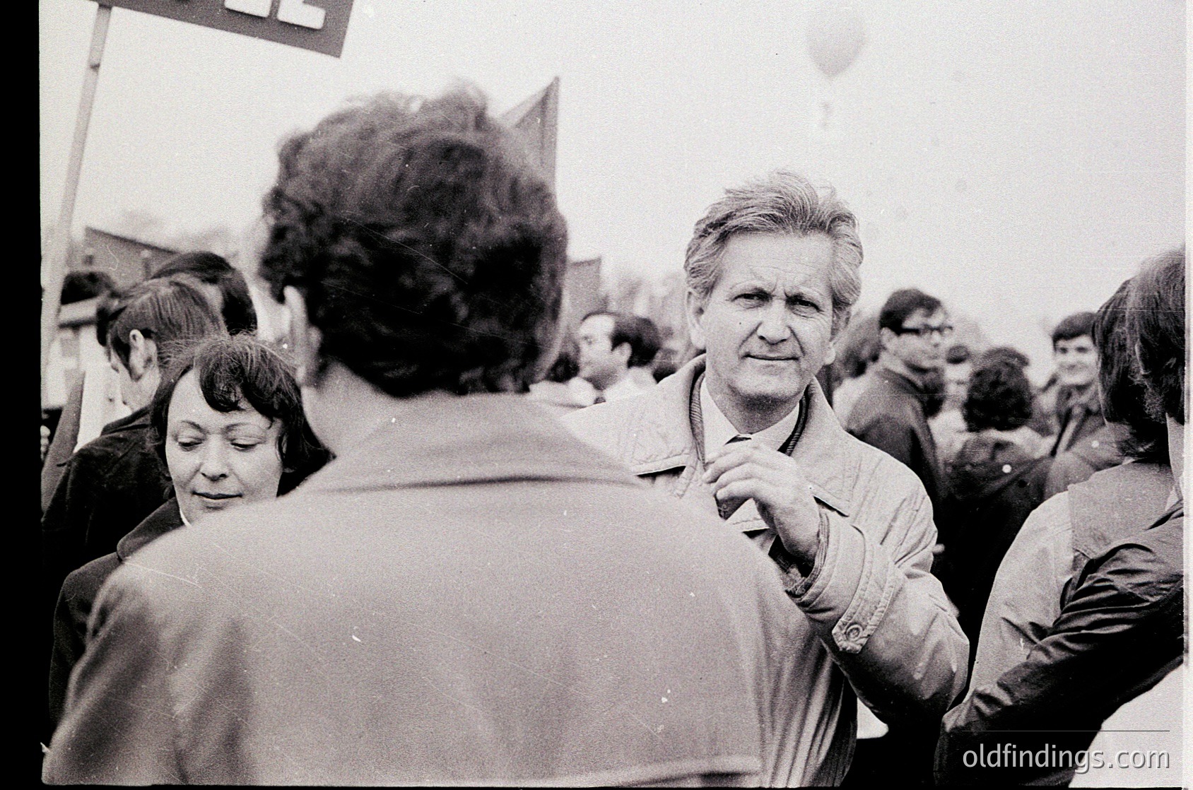 Black-and-white street protest with dense crowd, 1970s–1980s Eastern Bloc. Central figure in jacket gestures toward camera, surrounded by blurred faces and a visible sign. Urban setting with indistinct buildings and overcast sky. Captures collective activism and public demonstration.
