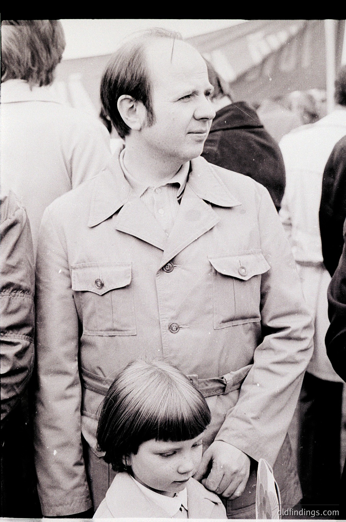 Black-and-white portrait of a man in a button-up shirt with a child standing beside him, likely mid-1970s. The man’s hairstyle and clothing suggest a Western European or North American setting. Crowd and blurred background indicate an outdoor public event or gathering.