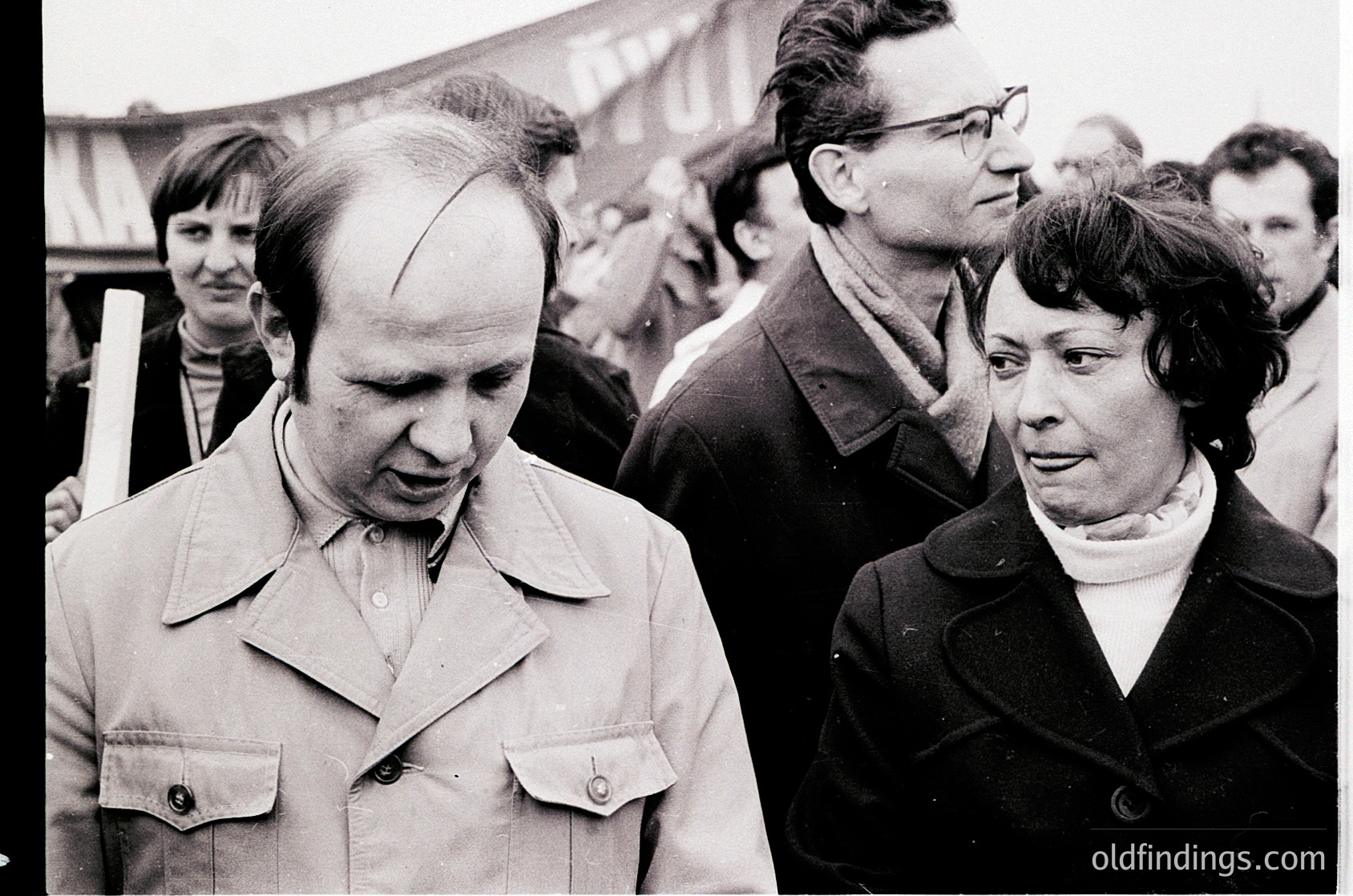 Black-and-white street scene from the 1960s–70s, featuring a man in a button-down shirt with a receding hairline and a woman in a collared coat, walking outdoors. Crowd and urban backdrop suggest a public demonstration or rally. Distinctive 1960s hairstyles and clothing styles ( ).
