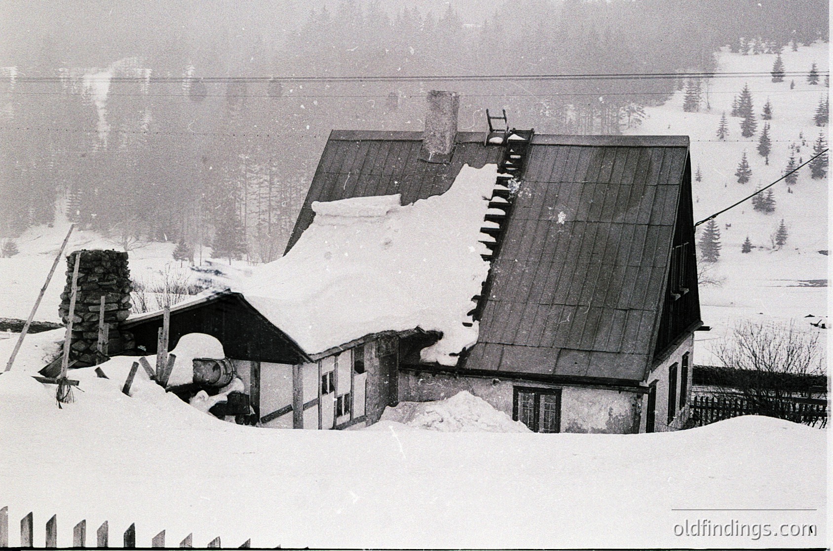 Snow-covered alpine cabin with steeply pitched roof, partially buried in winter. Rustic stone chimney and wooden exterior suggest traditional mountain architecture. Dense forest and misty valley in background. Likely Eastern European alpine region, mid-20th century.