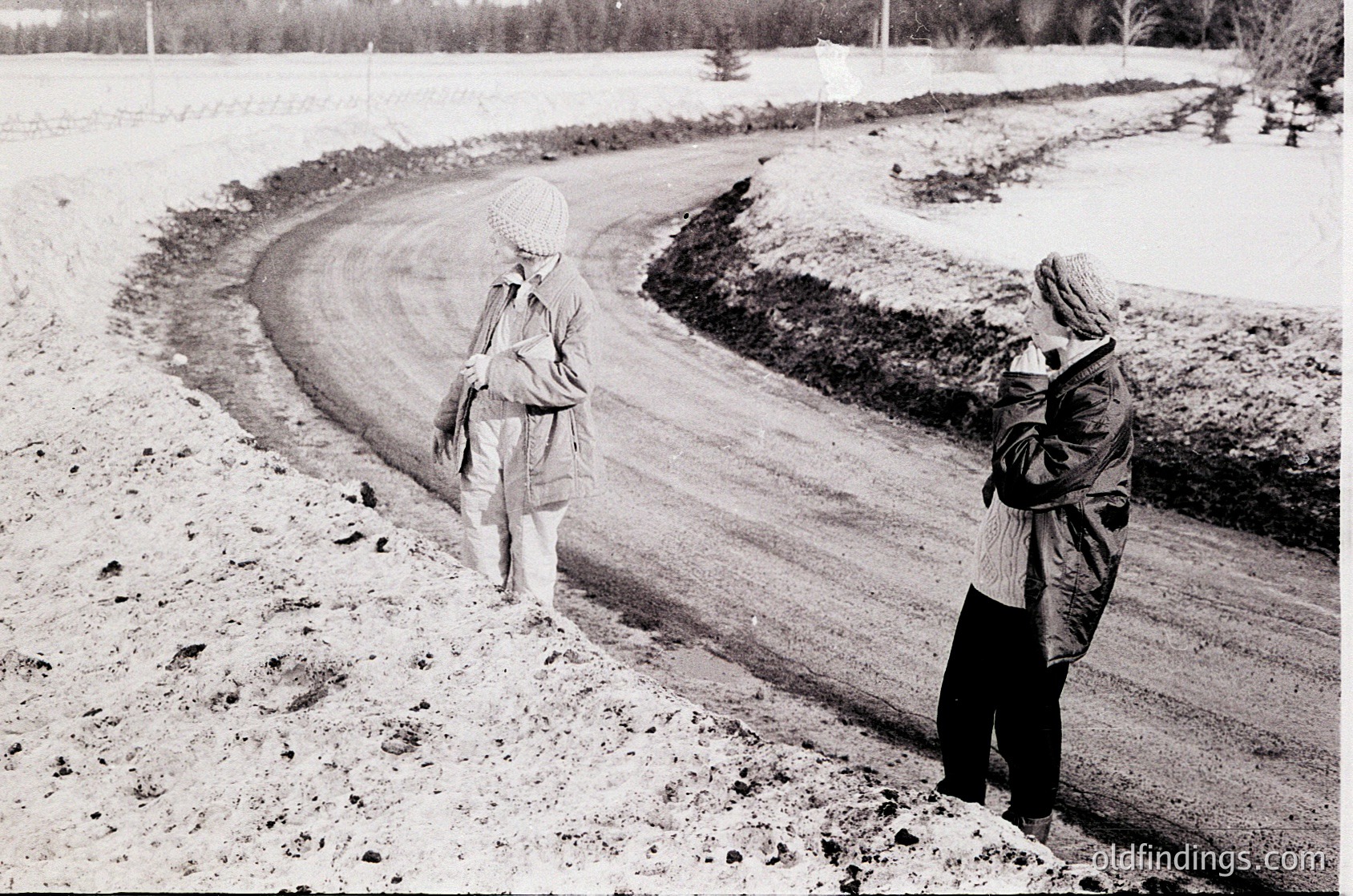 Mid-20th century black-and-white snapshot of two figures on a snow-covered rural road, likely . Adult holds child’s hands, both bundled in winter coats and hats. Snow blankets fields and roadside, with tire tracks visible. Evokes nostalgic and .