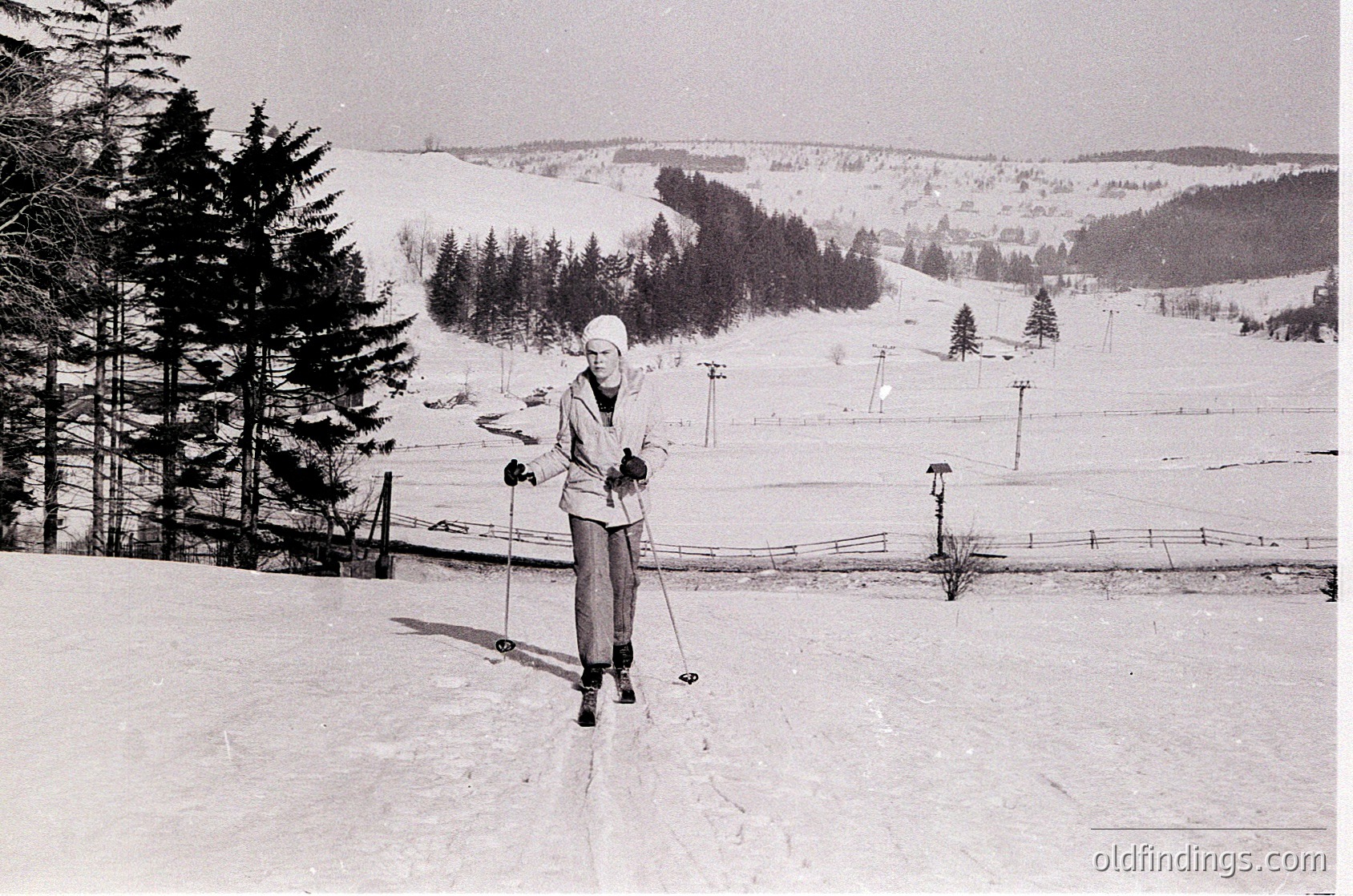 Mid-century cross-country skier in snowy forest, posing with poles. Classic 1960s winter attire—long-sleeve top, gloves, and hat. Snow-covered pine trees and fenced ski trail in background.