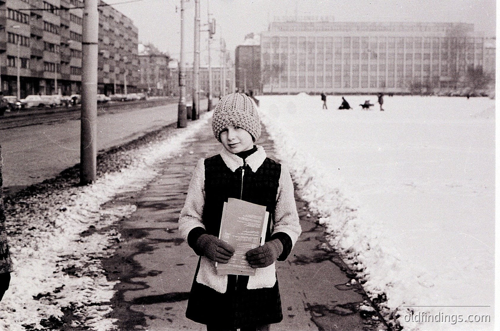 Winter street scene featuring a child in winter attire holding a book, standing on a snow-covered sidewalk beside a road. Mid-20th century urban setting with Soviet-era architecture and tram tracks.