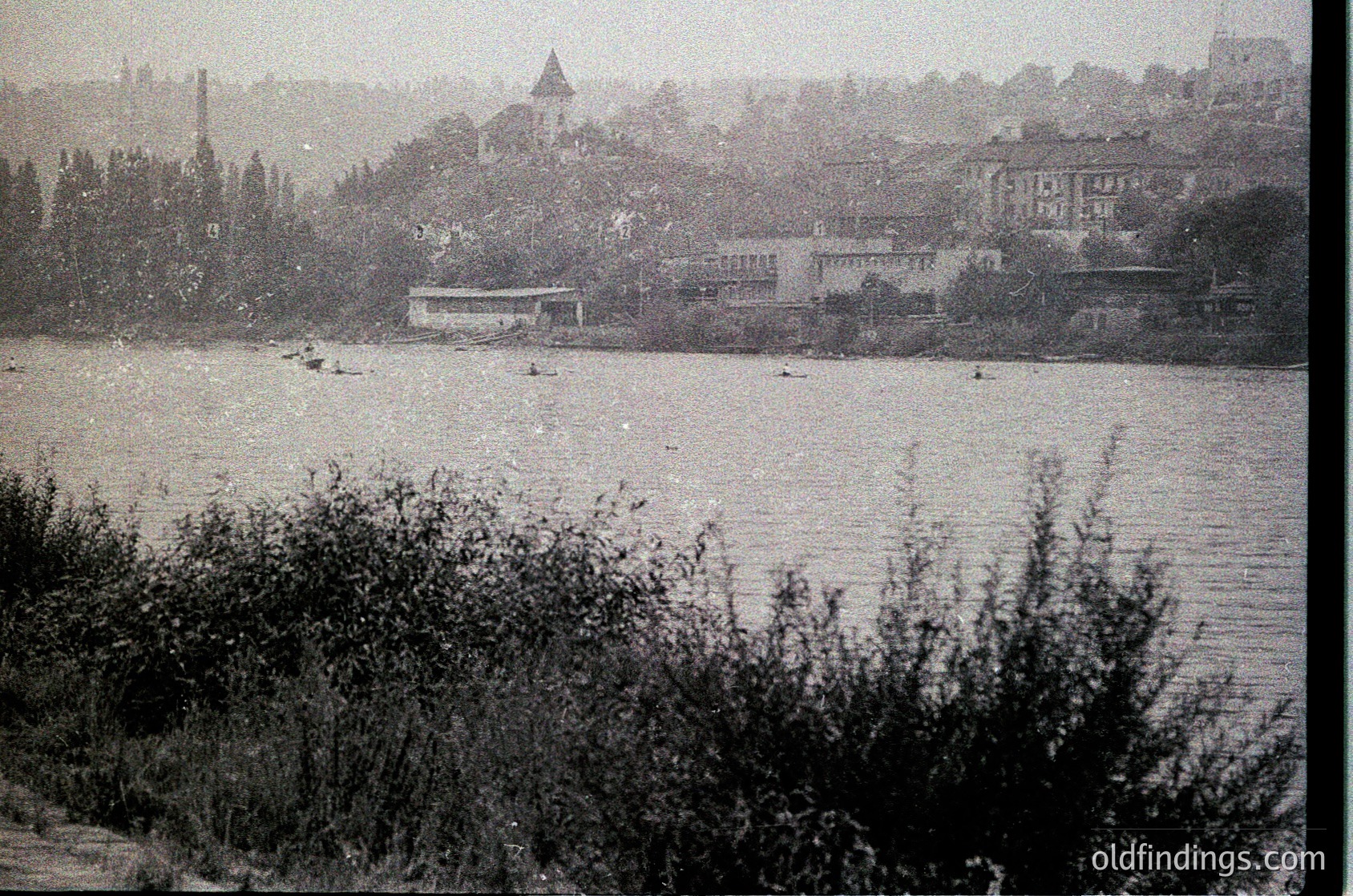 Vintage black-and-white riverside scene with dense foliage in foreground. Mid-20th century urban landscape featuring low-rise buildings, a prominent tower, and industrial chimneys. Waterfront area with small boats and a docked structure.