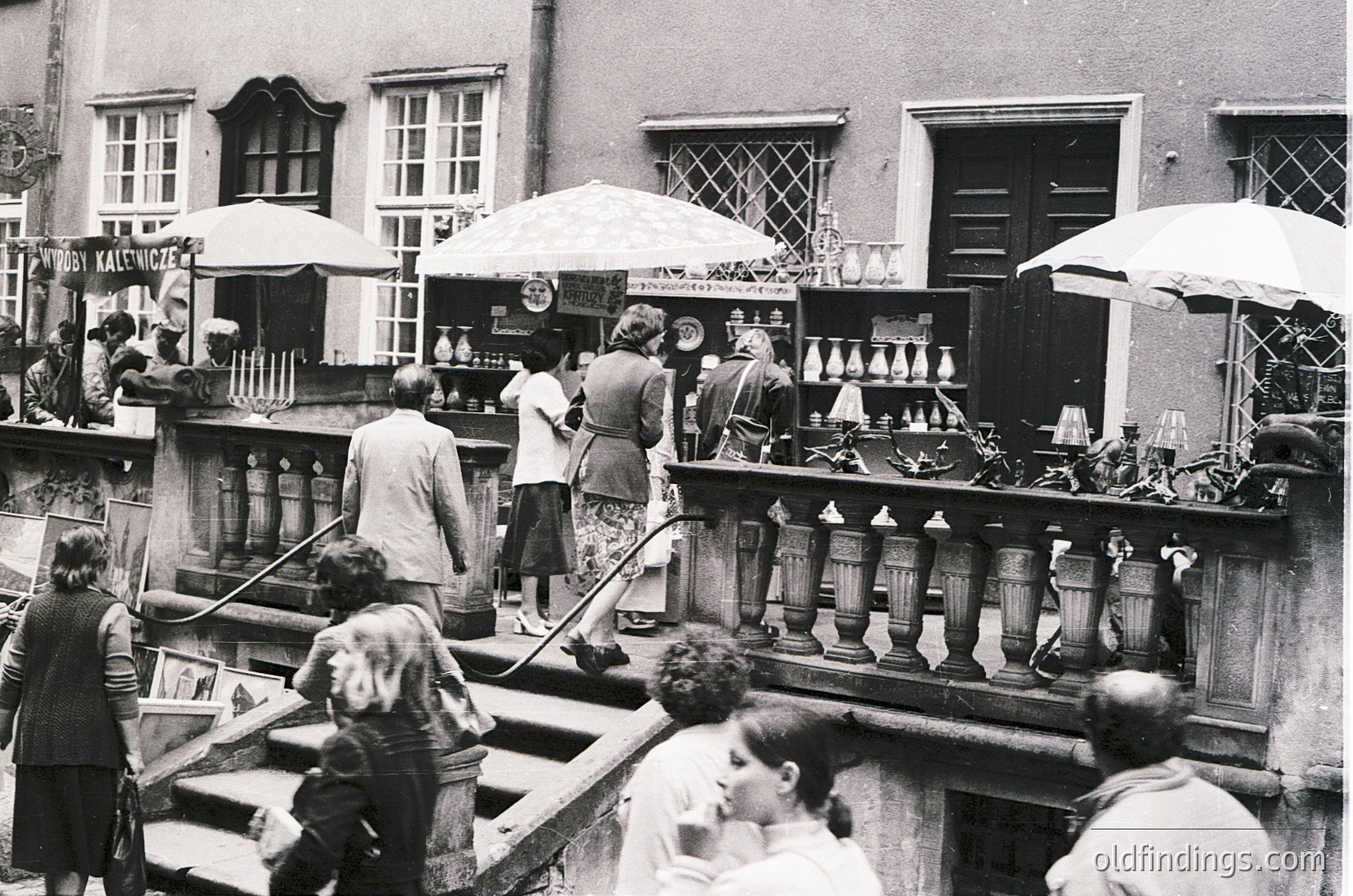 Vintage black-and-white courtyard market featuring brassware stalls under umbrellas. Crowd browsing ornate pitchers, teapots, and decorative items. Mid-20th century European street scene with multi-story residential/shop buildings.